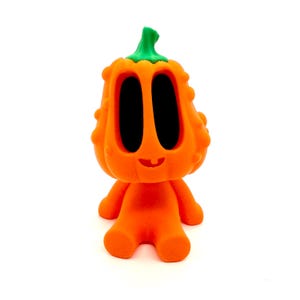 May include: A whimsical orange figurine resembling a pumpkin with large black eyes and a green stem. The figure has a smiling face and small arms and legs, perfect for Halloween or fall decor.