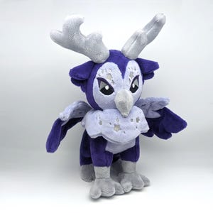 May include: A purple and grey plush toy with antlers, wings, and feet. The toy has a star pattern on its chest and a white beak.