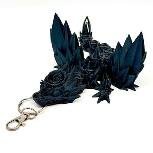 May include: A black 3D printed dragon keychain with a rose design. The dragon has wings and a long tail. The keychain is attached to a silver metal ring.