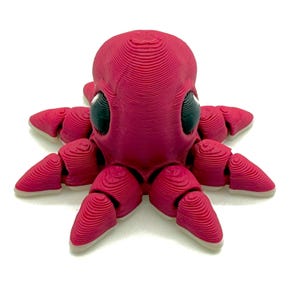May include: A 3D-printed octopus toy in a vibrant magenta color. The octopus has large, black eyes and articulated tentacles with a layered design. The toy is set against a white background.