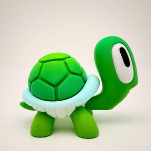 May include: A green and white turtle figurine with a textured shell and large, white eyes. The turtle is standing on a white surface.