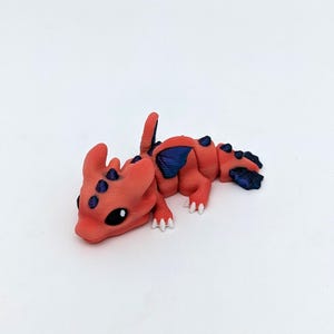 May include: A small, orange dragon figurine with blue accents on its wings and back. The dragon has white claws and black eyes. The figurine is lying down on a white surface.