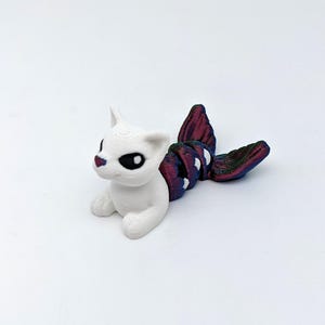 May include: A 3D-printed figurine of a cat with a mermaid tail. The cat is white with black eyes and a red nose. The tail is a gradient of purple, blue, and green. The cat is in a lying down position.