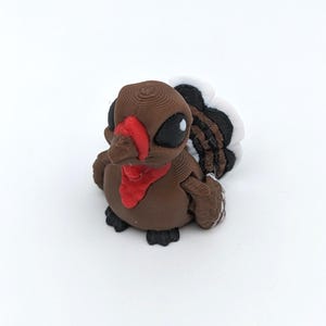 May include: A 3D-printed turkey figurine in brown, red, black, and white. The turkey has large black eyes, a red wattle, and a fan of feathers. The figurine is a cute, stylized representation of a turkey.