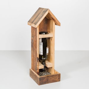 Könnte beinhalten: Ein rustikaler Vogelhaus-Futterspender in Form eines kleinen Hauses. Er enthält eine dunkle Weinflasche, mit einem kleinen Tablett für Samen am Boden. Das Holz hat ein natürliches, verwittertes Aussehen.