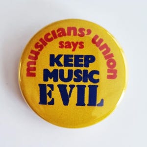 May include: A round, yellow button with the text "musicians' union says KEEP MUSIC EVIL" in red and blue lettering. The button has a glossy finish and a vintage aesthetic.