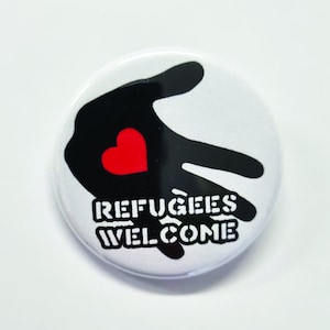 Puede incluir: Un botón blanco con una huella de mano negra y un corazón rojo. El texto "Refugees Welcome" está impreso en blanco en el botón.