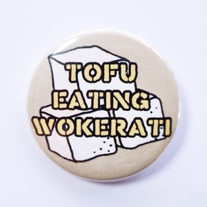 Może przedstawiać: Okrągła przypinka z beżowym tłem i żółto-czarnym napisem "TOFU EATING WOKERATI". Przypinka przedstawia grafikę trzech kostek tofu.