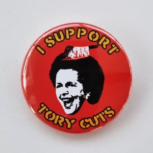 Puede incluir: Un botón rojo con el texto "I Support Tory Cuts" en letras amarillas. Una imagen en blanco y negro de una mujer con un cuchillo ensangrentado en la cabeza está en el centro del botón.
