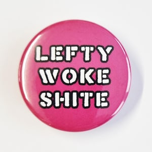 May include: A circular, cerise button featuring the text "LEFTY WOKE SHITE" in a bold, white, sans-serif font. The button has a shiny surface and is set against a white backdrop.