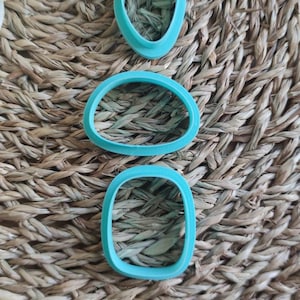 Op de afbeelding: Drie turquoise plastic koekjesvormen in verschillende vormen: een hart, een ovaal en een rechthoek. De vormen liggen op een geweven bruine achtergrond.