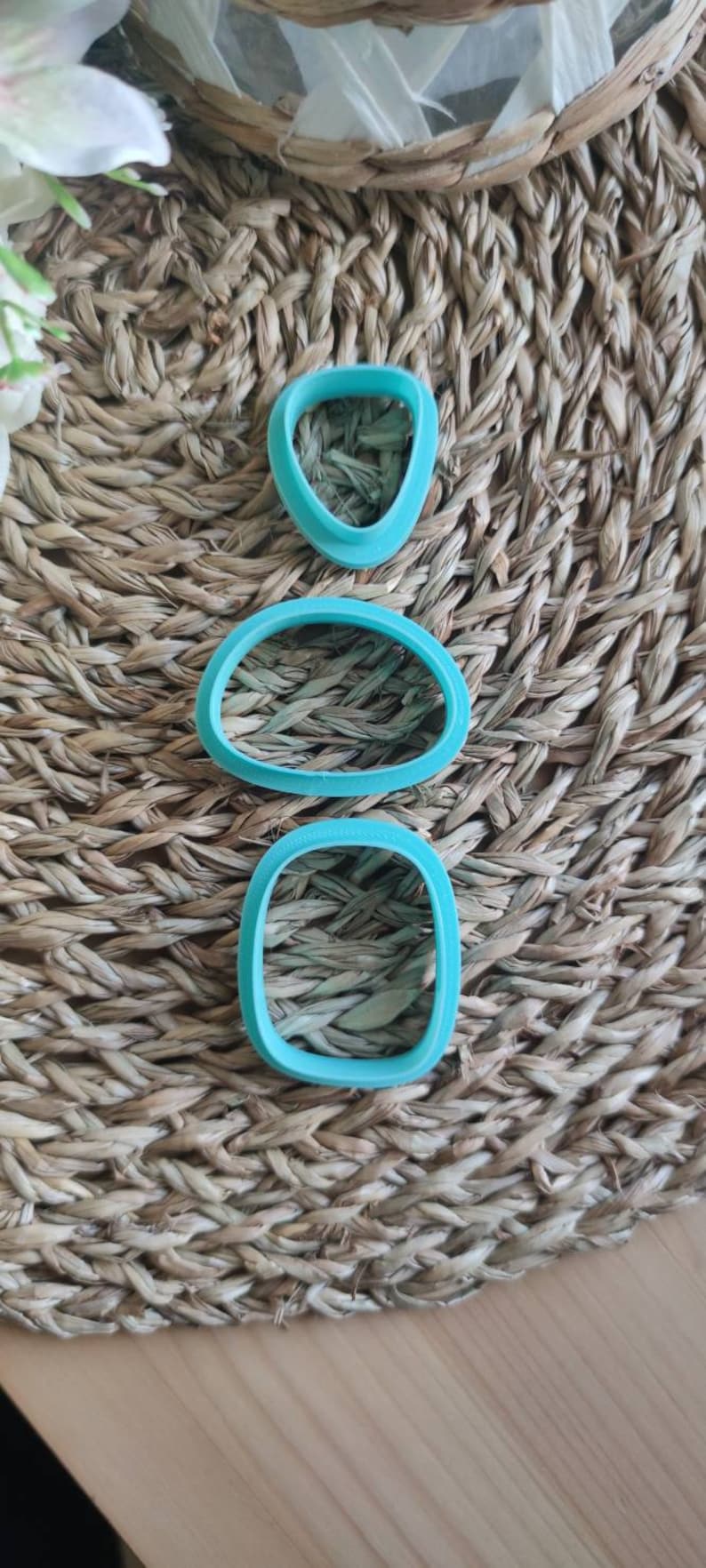 Op de afbeelding: Drie turquoise plastic koekjesvormen in verschillende vormen: een hart, een ovaal en een rechthoek. De vormen liggen op een geweven bruine achtergrond.