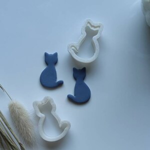 Op de afbeelding: Drie koekjesvormen in de vorm van een kat, twee zijn blauw en één is wit. De vormen liggen op een wit oppervlak.