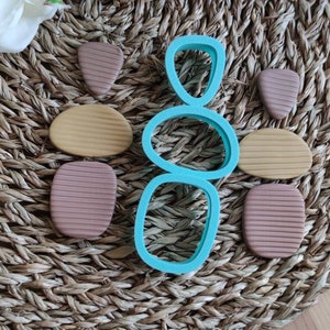 Op de afbeelding: Een set teal plastic koekjesvormen in de vorm van gestapelde stenen. De vormen liggen op een geweven bruine mat met bruine, beige en beige klei vormen.
