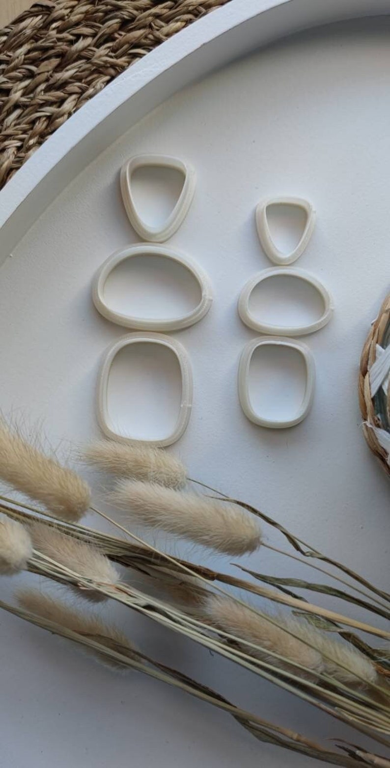 Op de afbeelding: Een set van vijf witte plastic koekjesvormen in verschillende vormen, waaronder een traan, een ovaal en een rechthoek, gerangschikt op een wit oppervlak.