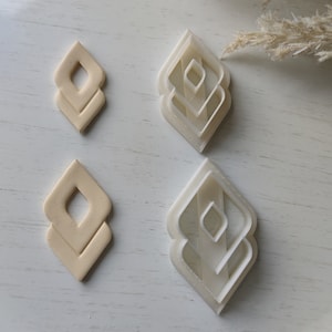 Op de afbeelding: Een bovenaanzicht toont vier geometrische oorbeluitstekers. Twee zijn beige en twee zijn wit. De uitstekers hebben een gelaagde, diamantachtige vorm. De achtergrond is een wit oppervlak met gedroogde, veervormige planten.