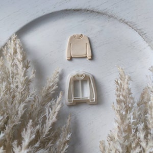 May include: A white plastic cookie cutter in the shape of a sweater with snowflake details. A beige plastic cookie cutter in the shape of a sweater with snowflake details is shown next to it.