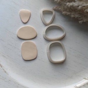 May include: A set of four polymer clay cutters in the shape of abstract organic shapes. The cutters are white and are arranged on a white surface. There are three beige polymer clay shapes next to the cutters.