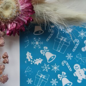 Pode incluir: Um estêncil azul com desenhos brancos temáticos de Natal, incluindo flocos de neve, sinos, bengalas de doces, presentes, renas e homens de gengibre.