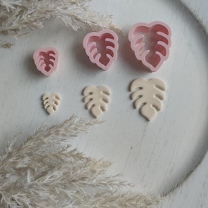 May include: A set of five pink and beige plastic cookie cutters shaped like monstera leaves. The cutters are arranged on a white surface with a fluffy white plant in the background.