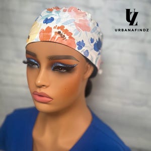 Op de afbeelding: Een witte scrubcap met een bloemenpatroon in tinten blauw, roze en oranje. De cap heeft een zwart elastiek en wordt gedragen door een persoon met blauwe oogschaduw.