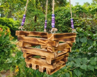 Puede incluir: Un macetero colgante de madera con forma cuadrada y acabado marrón rústico. El macetero está hecho de listones de madera apilados y tiene un diseño sencillo. Está suspendido por tres cuerdas con cuentas moradas y blancas.