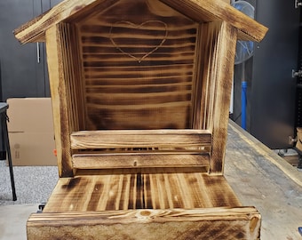 Puede incluir: Una casita de pájaros de madera con un corazón tallado en la parte delantera. La casita de pájaros está teñida de un color marrón oscuro y tiene un pequeño estante debajo.