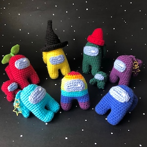 May include: A collection of colorful, crocheted Among Us characters. The characters are in various colors, including red, yellow, green, blue, and purple, and some have hats. The background is black with white star-like dots.