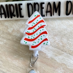 May include: A white Christmas tree-shaped retractable badge holder with red and green stripes and green polka dots. The holder is attached to a white retractable cord and a silver swivel clip.