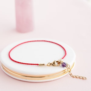 May include: A red string bracelet with a gold clasp and a small purple gemstone charm.