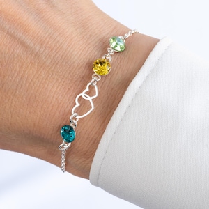 May include: A silver chain bracelet with three colourful crystal stones and a double heart charm. The stones are green, yellow, and teal.