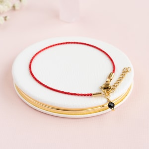 May include: A red cord bracelet with gold-colored clasps and a small black bead charm. The bracelet is displayed on a white, round surface with a gold rim, set against a soft pink background.