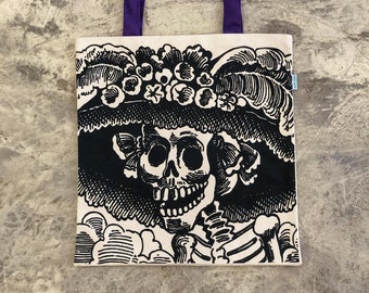 Op de afbeelding: Een crèmekleurige canvas tas met een zwarte inktillustratie van een skeletfiguur met een hoed met brede rand versierd met bloemen. De tas heeft twee paarse stoffen hengsels aan de bovenkant en een klein blauw label aan de zijkant.