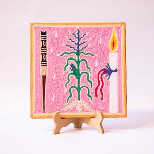 Puede incluir: Una obra de arte bordada en rosa con una planta verde central, una vela blanca con una llama amarilla y un objeto en blanco y negro. La obra de arte se exhibe en un pequeño caballete de madera.