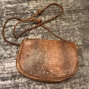May include: A brown, textured, semi-circular crossbody bag with a snakeskin-like pattern. The bag features a braided leather strap with two small, round, knotted leather accents and a single circular button closure on the front flap.