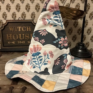 May include: A pointed hat featuring a white fabric base with a repeating pattern of dark gray bats and pink and blue floral sprigs. The wide brim is constructed from a quilted patchwork of square fabric panels in shades of blue, pink, and yellow.