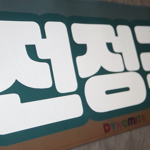 May include: A teal banner with white text that reads "전정국" in Korean. The text is in a bold, blocky font. The banner has the word "Dynamite" in rainbow letters at the bottom.
