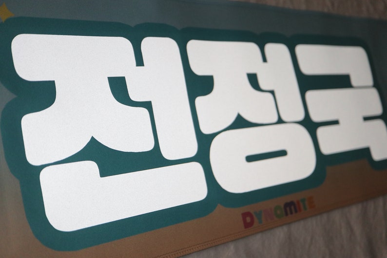 May include: A teal banner with white text that reads "전정국" in Korean. The text is in a bold, blocky font. The banner has the word "Dynamite" in rainbow letters at the bottom.