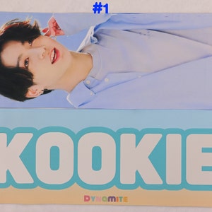 May include: A photo card featuring a portrait of a young man with black hair and a light blue shirt. The bottom of the card has a blue and white graphic with the word "KOOKIE" and the text "DYNAMITE" in smaller font.