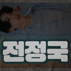 May include: A light blue and white fabric banner with the Korean text "전정국" in white, with the word "Dynamite" in rainbow letters below the text.