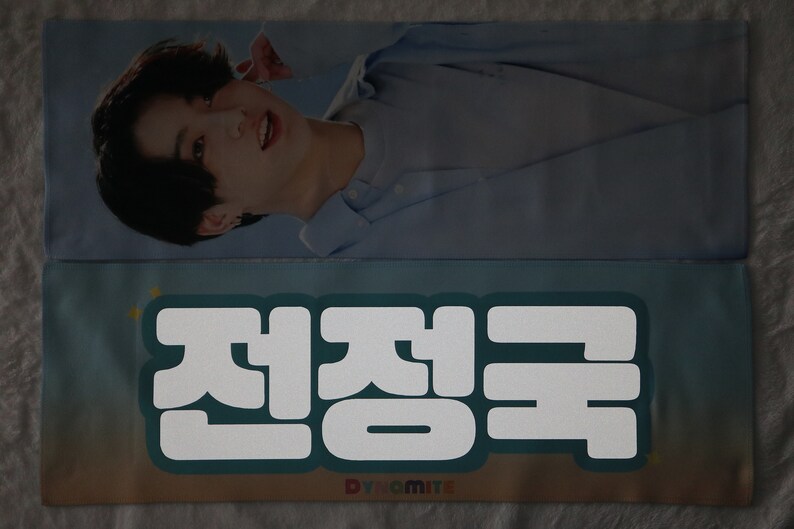 May include: A light blue and white fabric banner with the Korean text "전정국" in white, with the word "Dynamite" in rainbow letters below the text.