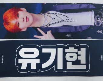 May include: A blue and white fabric banner with the text "MONSTA X" and "유기현" in white lettering. The banner features a photo of a person with orange hair wearing a black jacket and a silver necklace.