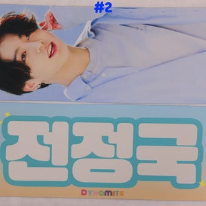 May include: A blue and white banner with the text "#2" in the top right corner. The bottom half of the banner features a large white text graphic that says "Dynamite" in English and "전정국" in Korean.