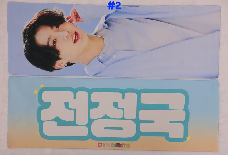 May include: A blue and white banner with the text "#2" in the top right corner. The bottom half of the banner features a large white text graphic that says "Dynamite" in English and "전정국" in Korean.