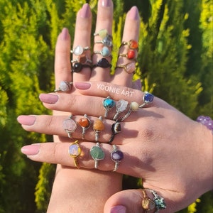 Puede incluir: Un maniquí de mano con una variedad de anillos con piedras preciosas de diferentes colores. Los anillos están hechos con alambre de plata y oro envuelto alrededor de las piedras. El texto "YOONIE ART" es visible en la mano.