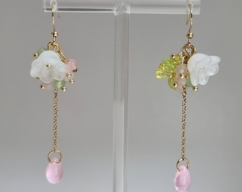 Op de afbeelding: Een paar gouden oorhangers met witte bloemencharms, roze en groene kralen en roze druppelvormige stenen aan de onderkant.