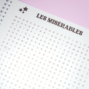 May include: A spiral-bound notebook with a word search puzzle. The title "LES MIS&Eacute;RABLES" is printed in a brown, stylized font above the puzzle grid. Two star graphics are in the upper left corner. The background is white with a pink border.