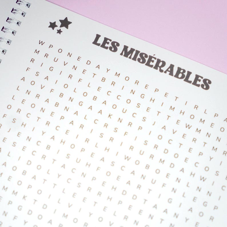 May include: A spiral-bound notebook with a word search puzzle. The title "LES MIS&Eacute;RABLES" is printed in a brown, stylized font above the puzzle grid. Two star graphics are in the upper left corner. The background is white with a pink border.