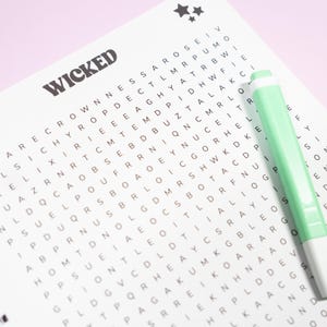 May include: A word search puzzle book with the word "WICKED" at the top. The puzzle is filled with letters and a light green pen is resting on the page. Two small star graphics are in the top right corner.