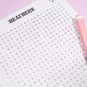 May include: A word search puzzle with the word "HEATHERS" at the top. The puzzle is on a white sheet of paper with a pink pen resting on the right side. The background is a light pink.
